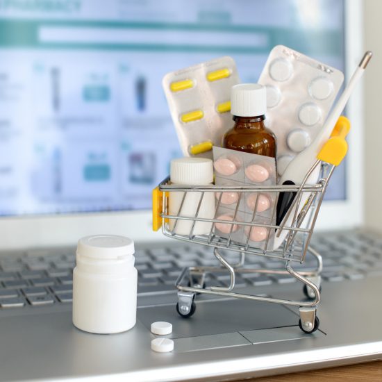 Shopping cart toy with medicines in front of laptop screen with pharmacy web site on it. Pills, blister packs, medical bottles, thermometer set. Health care and buying medications online.