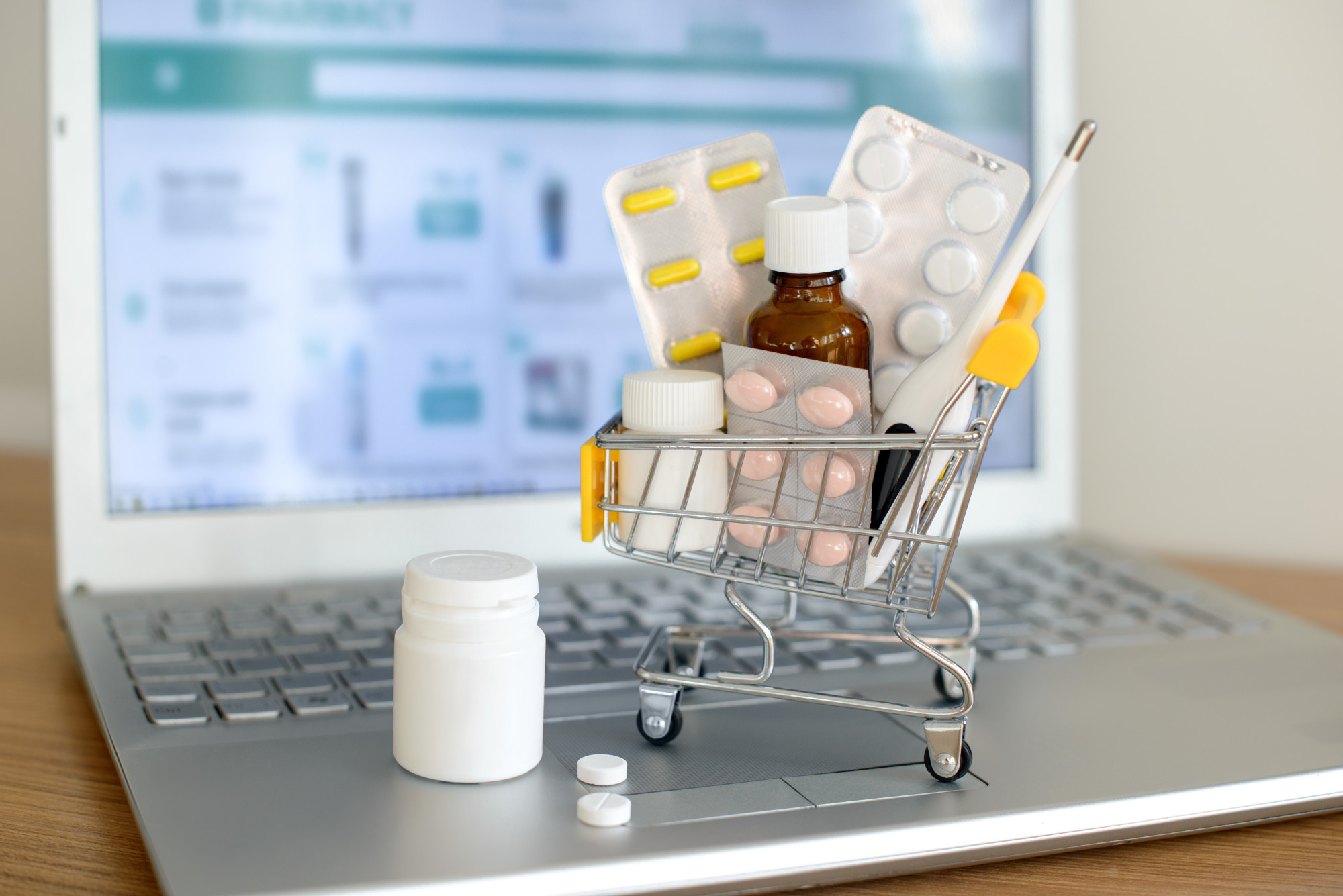 Shopping cart toy with medicines in front of laptop screen with pharmacy web site on it. Pills, blister packs, medical bottles, thermometer set. Health care and buying medications online.